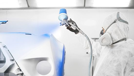 Male worker painting a car bumper in a white paint booth.